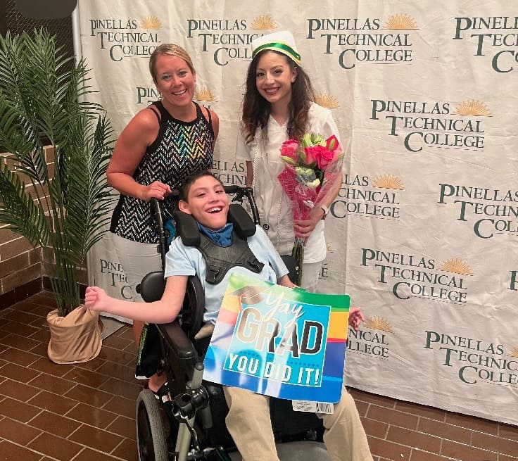 Two women smiling next to a pediatric homecare patient in a wheelchair who is holding a "congrats grad" sign