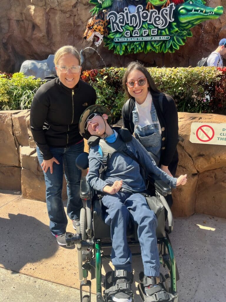 Two women in glasses standing behind a pediatric homecare patient, all are in front of the rainforest cafe sign