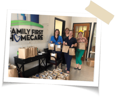 Four women holding up brown paper bags in front of a Family First Homecare Sign, there are also more brown paper bags lined up on the floor