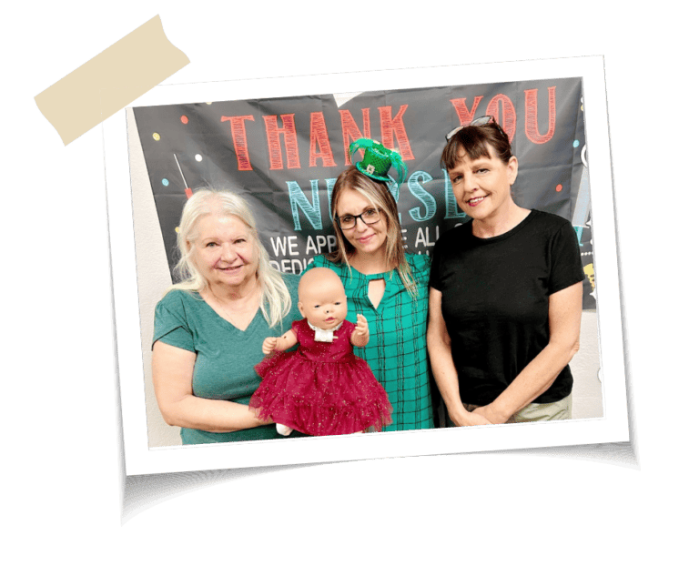 Two women in green and one woman in black hold up a baby doll wearing a red dress