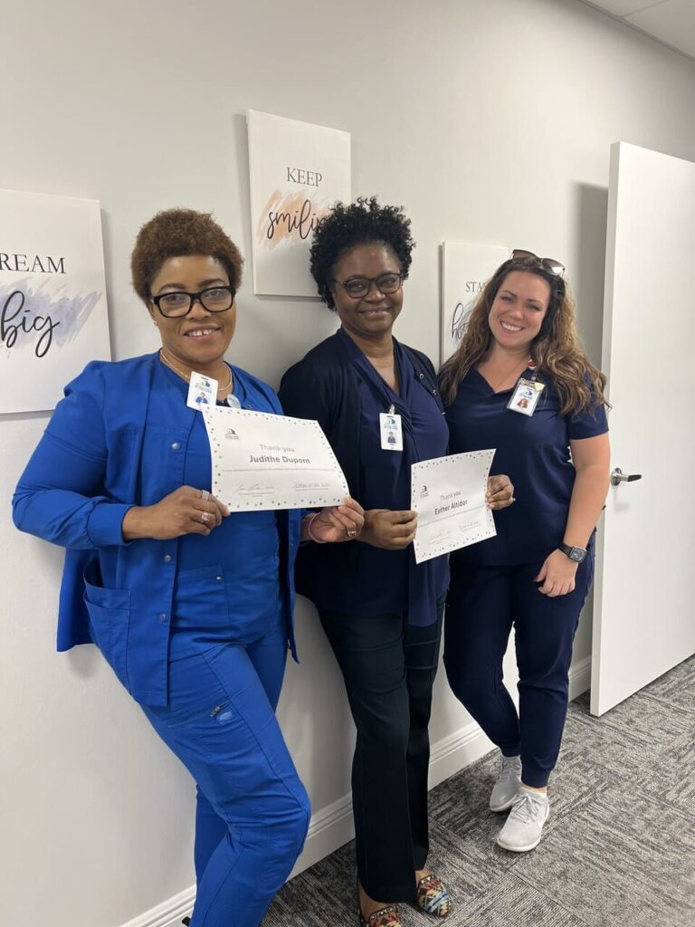 Three woman in blue scrubs smile at the camera, two are holding thank you certificates