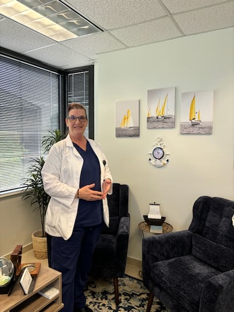 A woman in scrubs and a lab coat stands in an office with three sail boat pictures on the wall