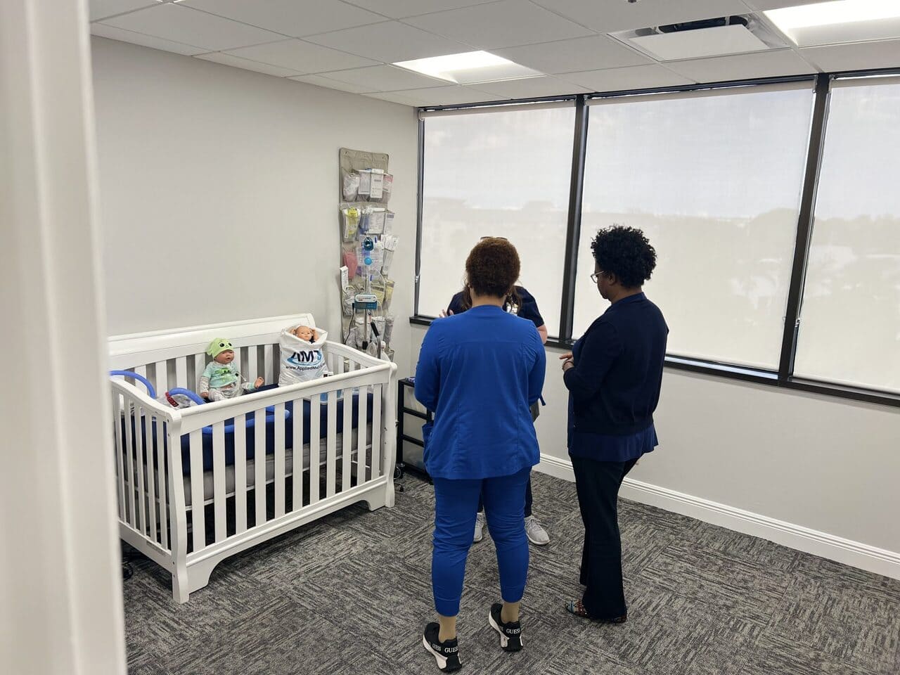 Three nurses in blue scrubs stand in a room near a crib with two baby dolls in the crib