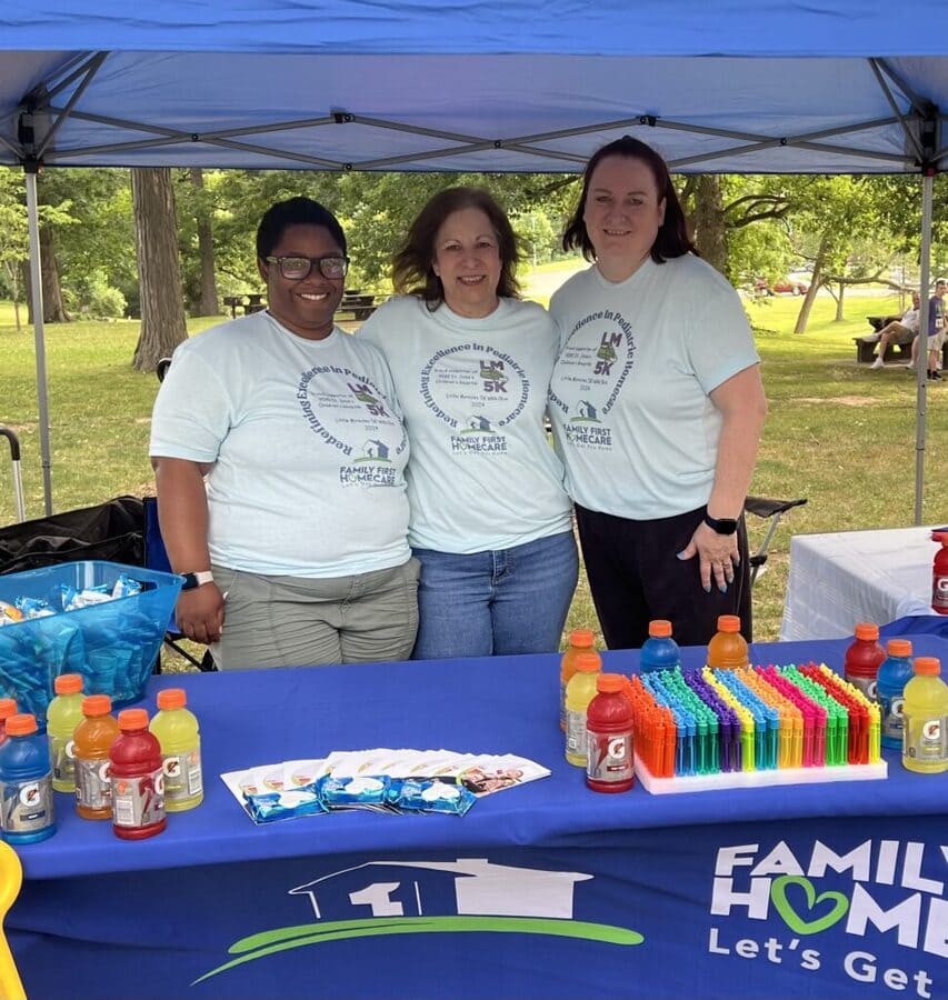 Three women standing in a Family First Homecare tent with Gatorade, bubbles and pamphlets 