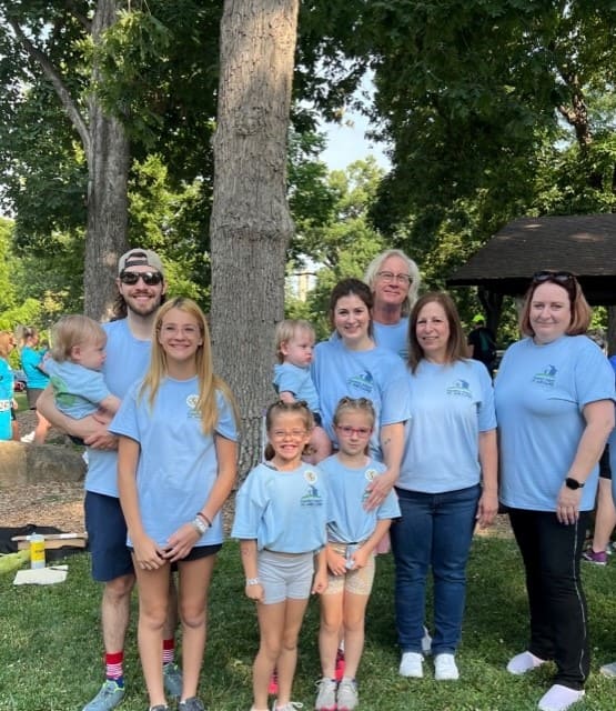 Adults and children in light blue shirts smiling at the camera in front of a tree