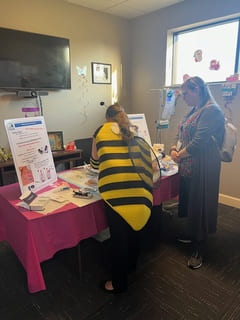 A woman in a bee costume stands next to a woman wearing grey, both are standing toward a Family First Homecare decorative table