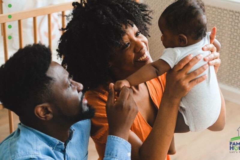 man sitting next to a woman holding a baby with a crib in the background