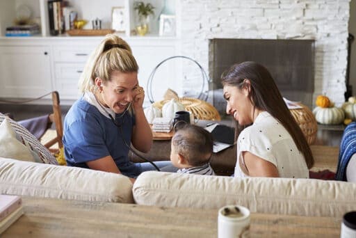 A nurse smiling while treating a pediatric homecare patient on a couch with the patient's mother sitting near